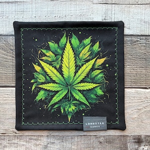May include: Black square fabric with a green and yellow cannabis leaf design. The leaf is centered on the fabric, surrounded by other leaves. The border is black with a green stitched edge. A small gray tag at the bottom right reads "LONESTAR HANKS".