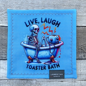 Pode incluir: Um tecido quadrado azul com uma borda costurada apresenta um esqueleto numa banheira com uma torradeira. O texto "LIVE, LAUGH, TOASTER BATH" está acima e abaixo da imagem. Uma pequena etiqueta na parte inferior diz "LONESTAR HANKS".