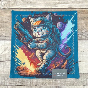 May include: Square blue handkerchief with a cartoon illustration of a cat astronaut holding a futuristic weapon. The cat is wearing a space helmet and suit, set against a cosmic background with planets and explosions. The text "LONESTAR HANKS" is visible.