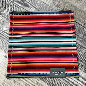May include: A square, multi-coloured fabric with horizontal stripes in red, orange, pink, green, blue, and black. The fabric has a white stitched border and a small grey label that reads "LONESTAR HANKS."