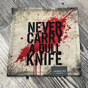 Può includere: Tessuto quadrato bianco sporco con la scritta "NEVER CARRY A DULL KNIFE" in nero, su un disegno a spruzzi rossi. Il tessuto ha un bordo cucito e una piccola etichetta con la scritta "LONESTAR HANKS".