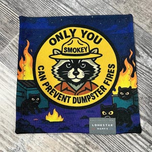 May include: Square fabric with a Smokey Bear design. The design features a raccoon wearing a ranger hat, surrounded by flames and the text "ONLY YOU CAN PREVENT DUMPSTER FIRES." Black cats and a dumpster are also visible. The fabric has a dark blue background.
