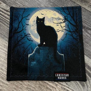 May include: A square fabric handkerchief featuring a black cat sitting on a tombstone against a large full moon, dark trees, and bats. The scene is rendered in shades of blue and black. A black tag with the white text LONESTAR HANKS is attached.