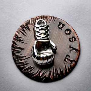 May include: A silver boxing glove charm is centered on a copper-colored, circular pendant. The word "Tyson" is stamped into the metal. The pendant has a textured, radial design around the edge.