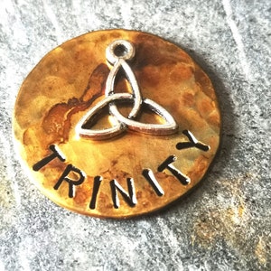 Puede incluir: Un amuleto redondo de metal de color dorado con la palabra "TRINITY" grabada en letras negras. Un dise&ntilde;o de nudo celta plateado est&aacute; centrado en el amuleto.