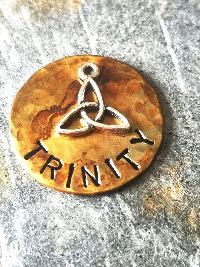 Puede incluir: Un amuleto redondo de metal de color dorado con la palabra "TRINITY" grabada en letras negras. Un dise&ntilde;o de nudo celta plateado est&aacute; centrado en el amuleto.