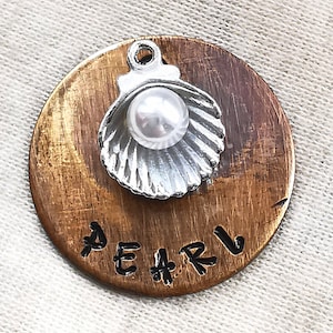 May include: A round, copper-coloured metal charm with the word "PEARL" engraved on it. A silver charm in the shape of a seashell with a white pearl inside is attached to the top.