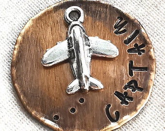May include: A copper-colored round charm with a silver airplane charm attached. The charm has the words "Captain" and "With" engraved on it.