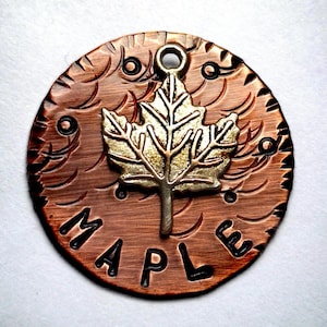 May include: A copper-colored round pendant with a textured surface and the word "MAPLE" embossed on it. A silver-colored maple leaf charm is attached to the pendant.
