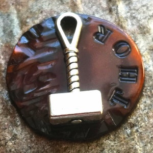 May include: A silver-colored hammer charm with a looped handle, attached to a round, brown, and black patterned disc with the word "Thor" embossed in black.