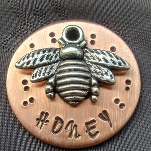 May include: A round copper charm featuring a silver bee design and the text "#HONEY" engraved on the front.