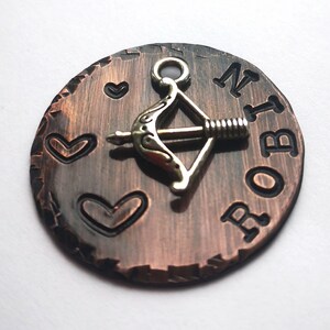 May include: A copper-colored round charm with a silver bow and arrow charm attached. The charm has the name "ROBIN" engraved on it and three heart shapes.