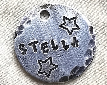 May include: A round, silver-colored pet tag with the name "STELLA" stamped in bold letters. Two star shapes and a flower are also stamped on the tag. The tag has a hammered texture and a hole for attachment.