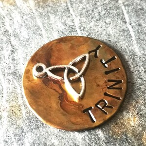 Puede incluir: Un amuleto redondo de color lat&oacute;n con un dise&ntilde;o de nudo celta plateado y la palabra "TRINITY" grabada alrededor del borde.
