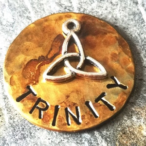 Puede incluir: Un disco de metal dorado con la palabra "TRINITY" grabada en letras negras. Un amuleto de nudo celta plateado est&aacute; centrado en el disco.