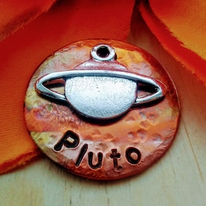 May include: A round, copper-colored pendant with a silver-colored planet design and the word "Pluto" etched below. The pendant has a hammered texture and a small loop at the top for hanging. The background is a vibrant orange.