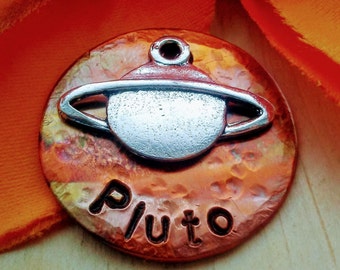 May include: A circular, copper-toned pendant featuring a silver planet design and the word "Pluto" etched beneath. The pendant has a hammered texture and a small loop for suspension. The background is a vivid orange.