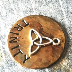 Puede incluir: Un colgante redondo de color lat&oacute;n con la palabra "TRINITY" grabada en el borde. Un dise&ntilde;o de nudo celta plateado se encuentra en el centro del colgante.