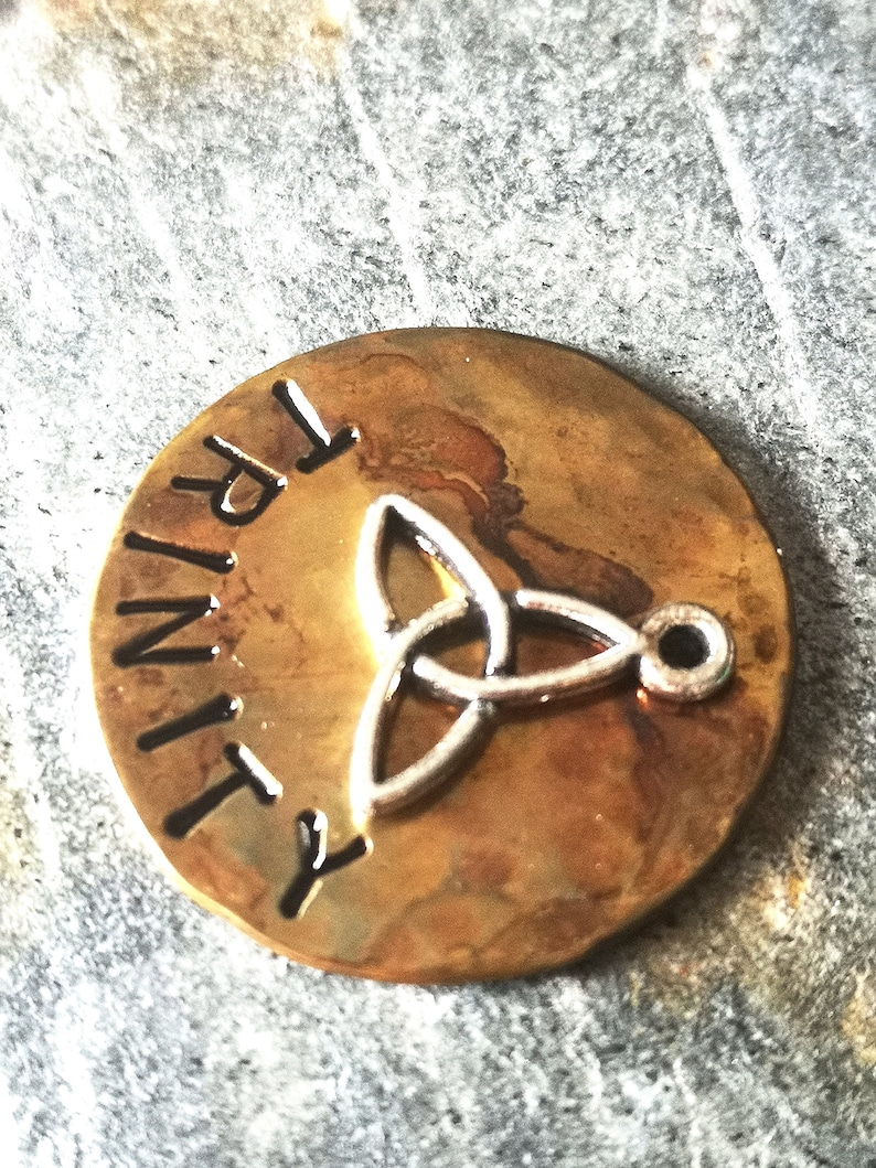 Puede incluir: Un colgante redondo de color lat&oacute;n con la palabra "TRINITY" grabada en el borde. Un dise&ntilde;o de nudo celta plateado se encuentra en el centro del colgante.
