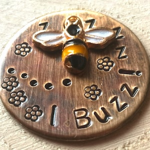 May include: A round, copper-coloured token with a small, gold-coloured bee charm in the centre. The bee has black and yellow stripes and white wings. The token is engraved with the words "I Buzz" and a pattern of small flowers.