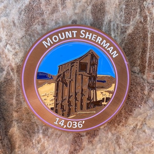 May include: A round, copper-colored enamel pin with a purple border. The pin features a blue sky, a brown building, and the text "MOUNT SHERMAN 14,036'" in white.