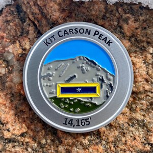 May include: A silver-colored round metal coin with a blue and white image of a mountain peak. The coin has the text "KIT CARSON PEAK" and "14,165'" on it.