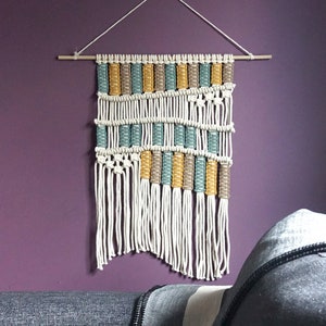 May include: A macrame wall hanging with a geometric pattern in white, yellow, and green yarn. The hanging is made with a wooden dowel and has a fringe at the bottom.
