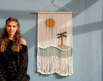 May include: A woman stands in front of a blue wall with a macrame wall hanging featuring a sun, palm tree, and waves. The wall hanging is made with white, blue, and yellow yarn.