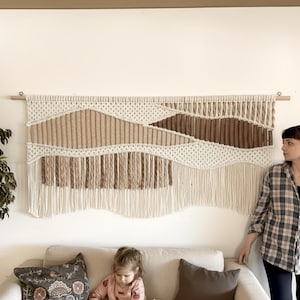 Peut inclure: Grande tenture murale en macramé aux tons neutres beige et marron. La pièce d'art textile présente des motifs géométriques et des franges, montée sur une cheville en bois. La décoration murale est exposée au-dessus d'un canapé beige.