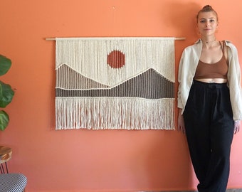 May include: A macrame wall hanging with a sunset scene featuring a large orange sun, brown mountains, and white and brown strands. The wall hanging is on a peach colored wall with a woman standing in front of it.