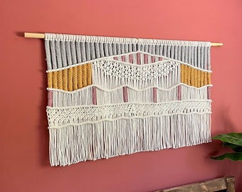 May include: A white, gray, pink, and yellow macrame wall hanging with a wooden dowel at the top. The wall hanging is made of knotted yarn and has a fringe at the bottom.