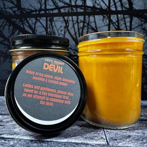 May include: Two glass jars with black lids. One jar is open, revealing a label with the text "LATE NIGHT DEVIL" and scent notes. The other jar is filled with a yellow substance. The jars are on a textured surface.