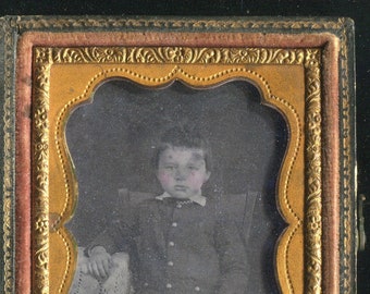 May include: A Victorian-era portrait of a child in a dark suit, seated in a chair. The image is framed in a decorative gold and red case. The child has rosy cheeks and a white collar.