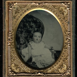 May include: A black and white antique photograph of a baby in a white dress, seated in a floral patterned chair. The oval image is framed with ornate gold detailing and encased in a rectangular frame.