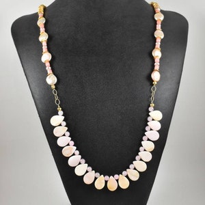 Puede incluir: Un delicado collar exhibido en un maniquí negro. El collar presenta una combinación de cuentas en forma de lágrima y redondas, de color rosa claro y crema. La cadena y los detalles dorados completan el diseño. Es una joya.