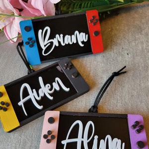 May include: Three wooden ornaments shaped like a Nintendo Switch console with different colored borders. The ornaments are personalized with the names Briana, Aiden, and Alina in white lettering.