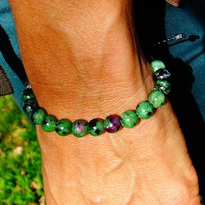 Peut inclure: Un bracelet en pierres précieuses vertes et roses avec une fermeture en cordon noir. Le bracelet est porté au poignet.