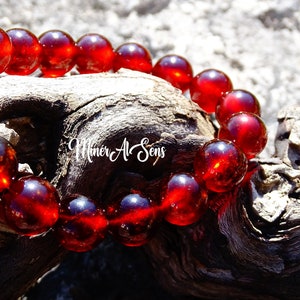 Puede incluir: Un brazalete de piedras preciosas rojas con cuentas redondas. El brazalete está hecho de piedras rojas pulidas y translúcidas. El brazalete está sobre un trozo de madera flotante.