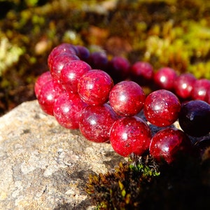 May include: A red gemstone bracelet with round beads. The bracelet is on a rock with green moss.