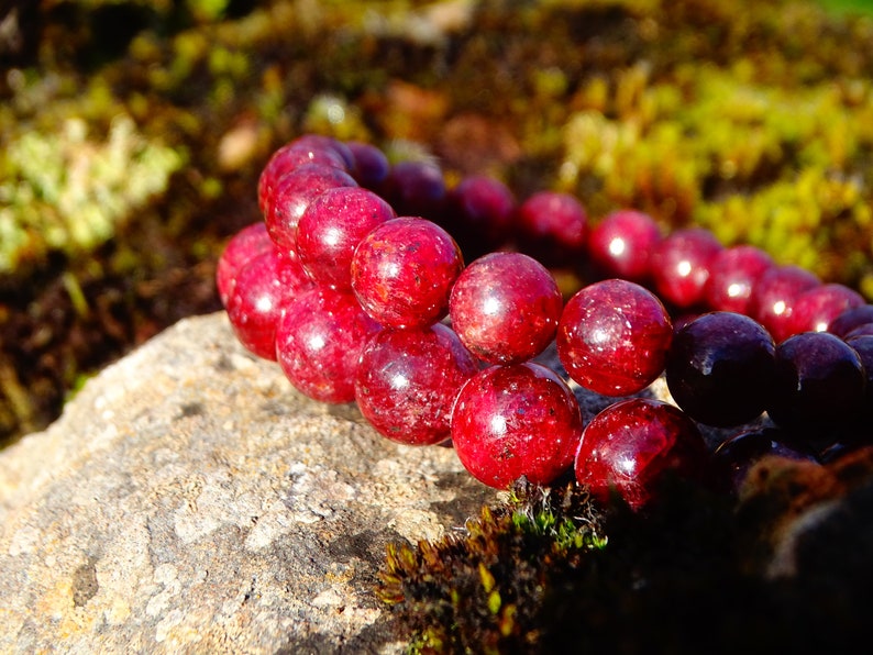 May include: A red gemstone bracelet with round beads. The bracelet is on a rock with green moss.