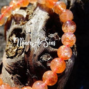 Puede incluir: Un primer plano de un brazalete de piedra de sol con cuentas de color naranja y rojo. El brazalete está sobre un trozo de madera con el texto "MINERAL & SENS" visible.