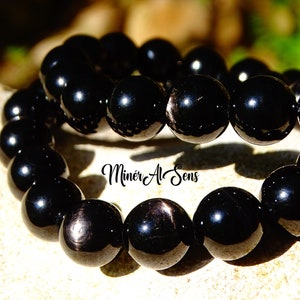 Puede incluir: Pulsera de cuentas de obsidiana negra con acabado pulido. Las cuentas son redondas y tienen un ligero brillo. La pulsera está apilada sobre sí misma, mostrando las cuentas desde múltiples ángulos.