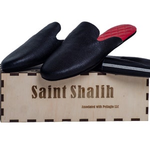 May include: Black leather slip-on shoes with red quilted insoles rest on a wooden box. The box is engraved with "Saint Shalih" and "Associated with Pellagio LLC". A black shoe bag with a silver stripe is also present.