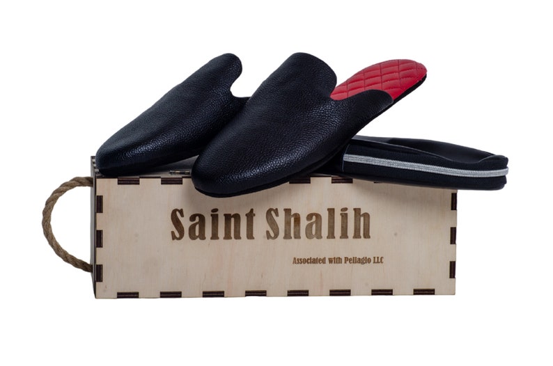May include: Black leather slip-on shoes with red quilted insoles rest on a wooden box. The box is engraved with "Saint Shalih" and "Associated with Pellagio LLC". A black shoe bag with a silver stripe is also present.