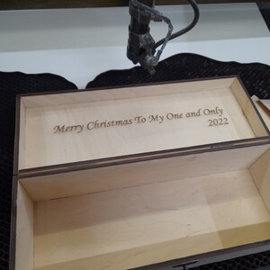 May include: A wooden box with a hinged lid. The lid is open and the inside of the box is visible. The lid is engraved with the text "Merry Christmas To My One and Only 2022".