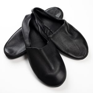 May include: Black leather ballet slippers with a soft, rounded toe. The slippers are shown with a matching black zippered case. The leather has a slight sheen, and the design is simple and elegant.