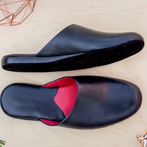Puede incluir: Dos zapatillas de cuero negro con forro rojo se muestran sobre una superficie de madera clara. Una es abierta, la otra cerrada. Objetos decorativos de color cobre en el fondo completan la presentaci&oacute;n.
