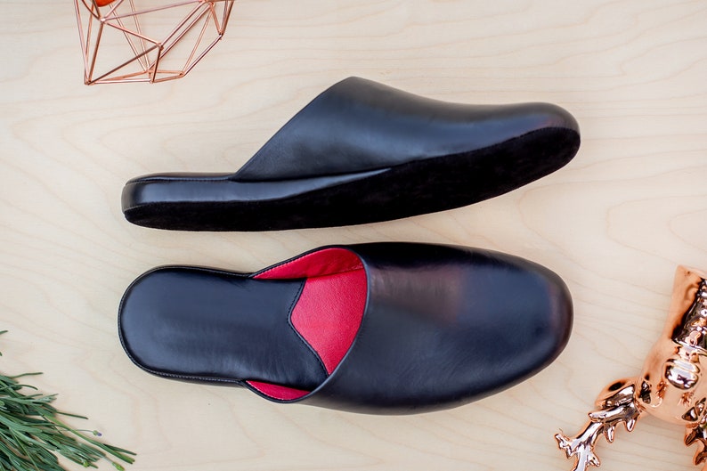 Puede incluir: Dos zapatillas de cuero negro con forro rojo se muestran sobre una superficie de madera clara. Una es abierta, la otra cerrada. Objetos decorativos de color cobre en el fondo completan la presentaci&oacute;n.
