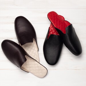 May include: Two pairs of leather slippers. One pair is black with red quilted lining, the other is dark brown with cream quilted lining. The slippers are on a white wooden surface.