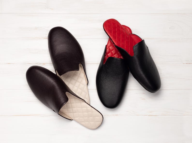 May include: Two pairs of leather slippers. One pair is black with red quilted lining, the other is dark brown with cream quilted lining. The slippers are on a white wooden surface.